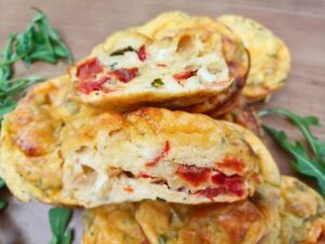 Close up of a cut open cottage cheese egg bite or muffin on top of other savory muffins on a wooden board with green garnish in the background