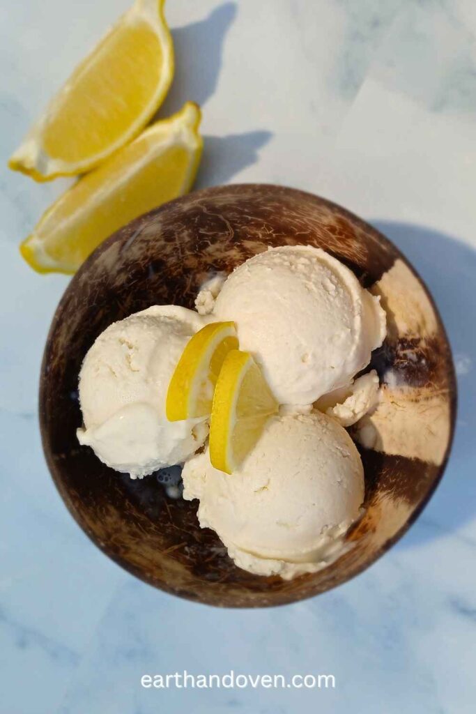 Three scoops of lemon flavored ice cream in a brown coconut shell bowl with wedges of lemon on the side and on top