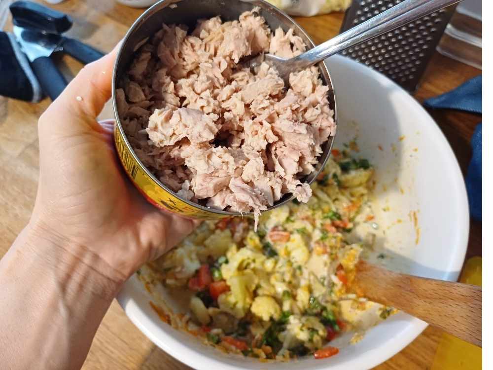 A hand holding a large can of tuna and the other hand is taking out the tuna to add to a larger bowl with vegetables