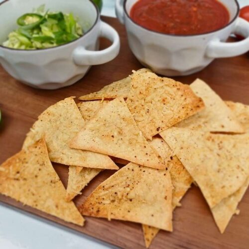 Keto tortilla chips on a wooden board with guacamole and salasa in the background