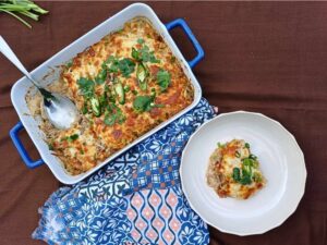 Top down view of a keto taco Mexican style caserole in a blue regtangular baking dish with a serving in a bowl on a brown tablecloth with a colorful towel on the table