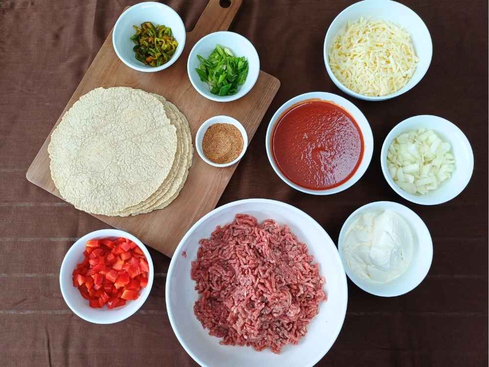 Ingredients in white bowls prepared for keto beef echilada casserole including ground beef, tortillas, sour cream and chopped peppers
