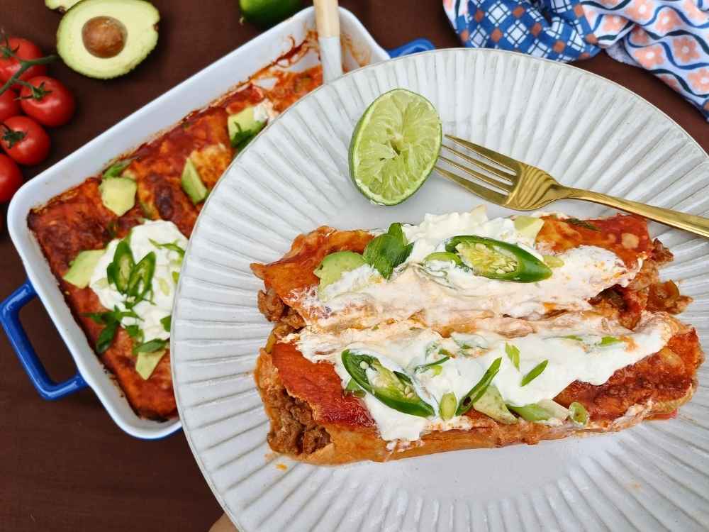 A serving of beef enchilada casserole presented on a white plate with sourcream and chillis on top and a gold fork, a wedge on lime on the side