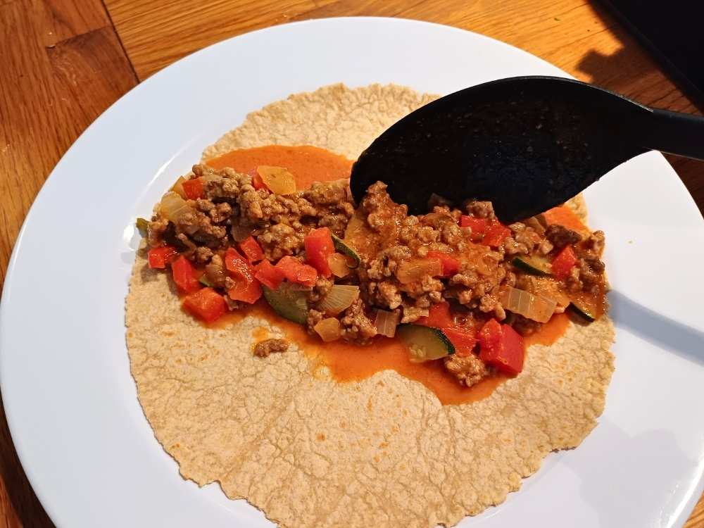 Ground beef enchilada mixture is being added to the tortilla with a spoon