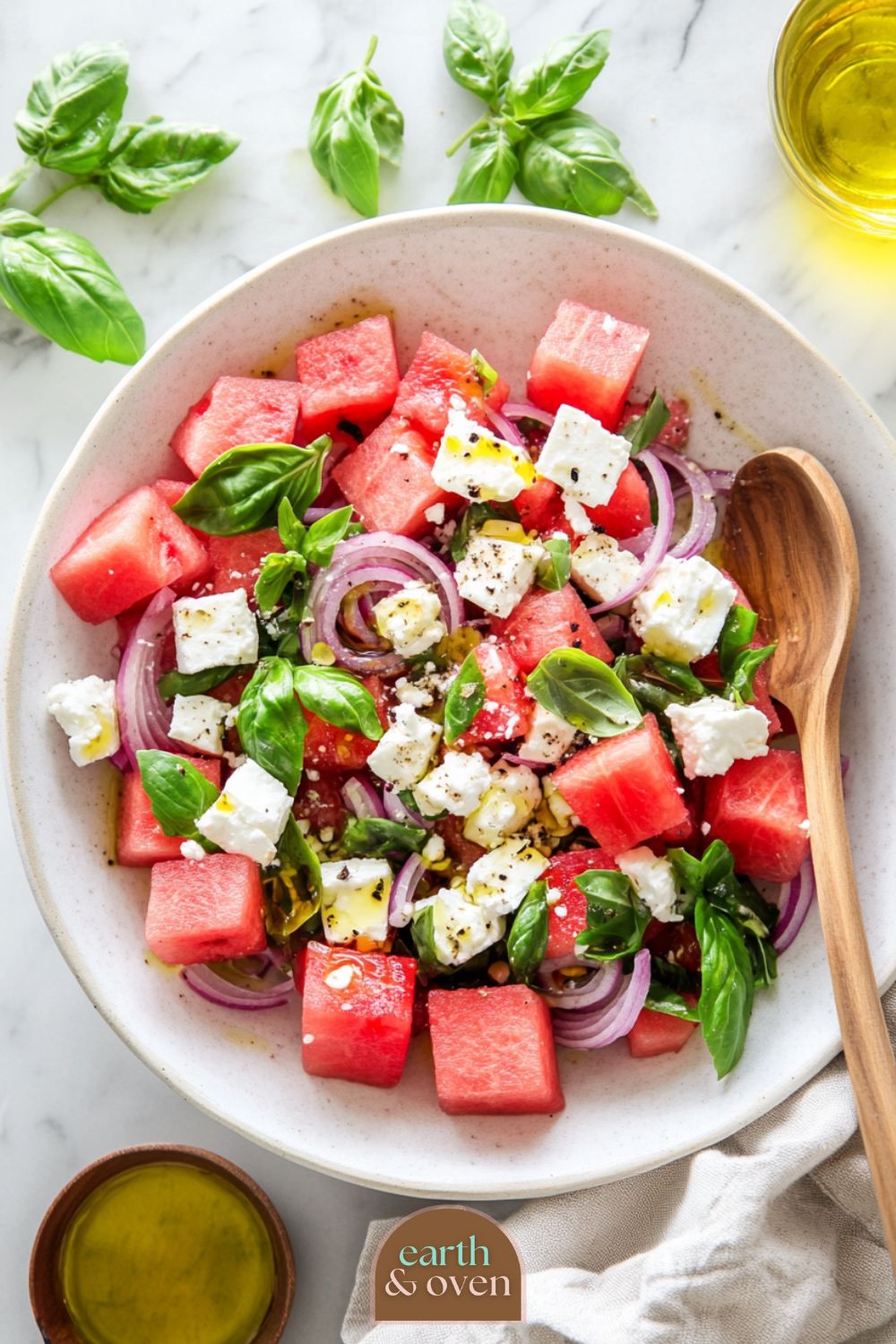 Fresh watermelon salad with feta, basil, spanish onions arranged in a white ceramic bowl and a wooden spoon on a marble benchtop with olive oil dressing on the salad and on the side.