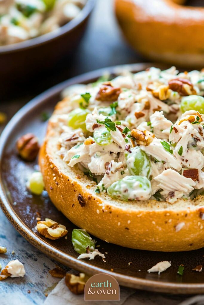 Sourdough bagel bottom half topped with creamy chciken salad with grapes and wallnuts on a brown plate.