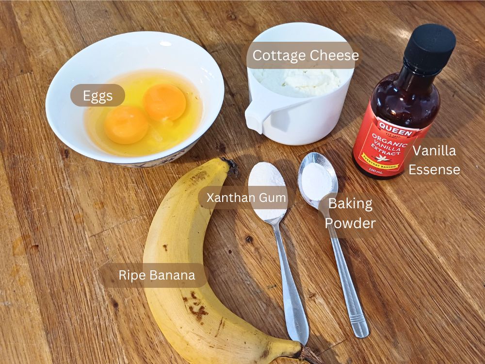 Ingredients displayed on a wooden bench including banana, cracked eggs in a white bowl, cottage cheese and other flavorings, clearly labeled.