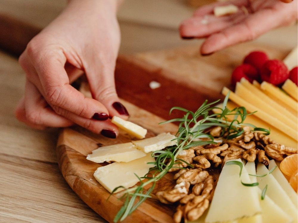 A womans hand visible assembling a summer charcuterie board, holding a piece of cheese which is about to be placed on a board next to wallnuts. Other grazing board items visible on a round wooden board.