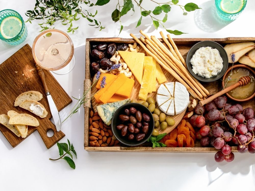 A wooden tray board filled with breakfast grazing items like cheeses, yogurt, bread sticks, olives and ham.