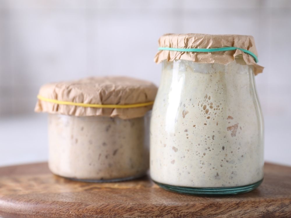 Sourdough discard in 2 glass jars with brown paper cover on a wooden board