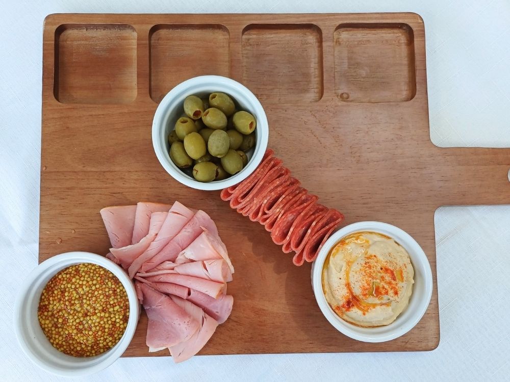 A wooden board with three ramekins filled with olives, mustard and hummus and some ham and salami folded in between.