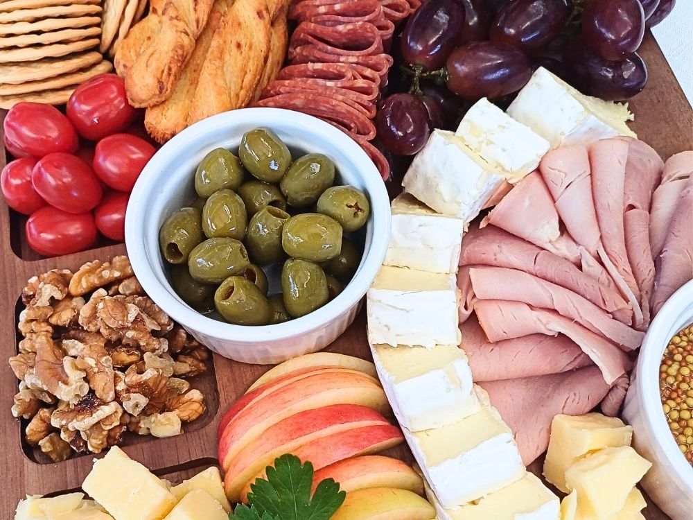 Close up of a simple charuterie board with ham, cheeses, slami, olives and fruits.