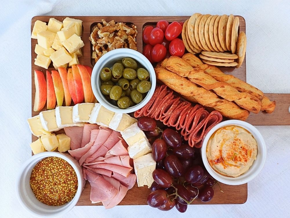 Top down view of a simple charcuterie board with ham, salami, crackers, cheeses, wallnuts, olives and hummus.