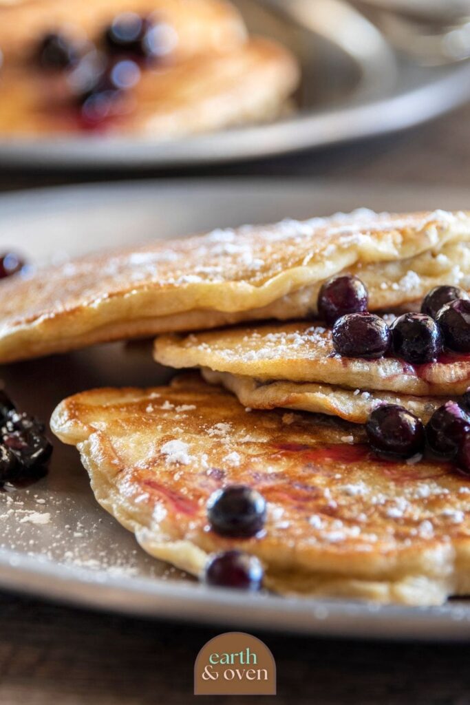 Stack of soudrough discard pancakes drizzled with maple syrup and topped with fresh blueberries with a bit of icing sugar close up