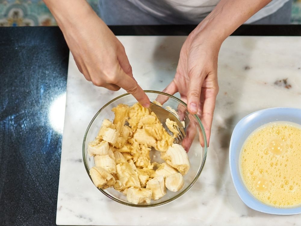 Top view of woman's hands mashing bananas in a glass bowl with a fork.