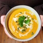 A woman's hands holding a bowl of warm homemade butternut squash soup sprnkled with pumpkin seeds. Woman wearing a brown sweater.