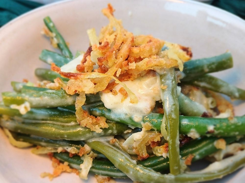 A white ceramic bowl filled with a serving of saucy green bean casserole topped with crispy French onions.