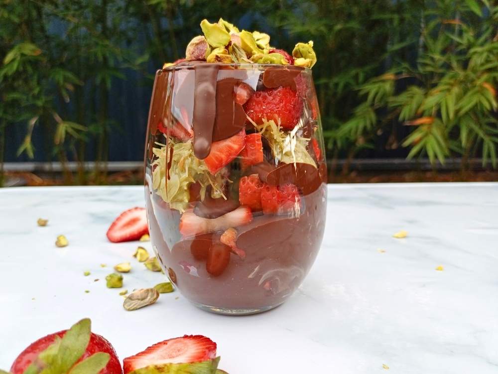 A glass cup filled with melted chocolate, chopped strawberries, kataifi pastry and pistachio cream