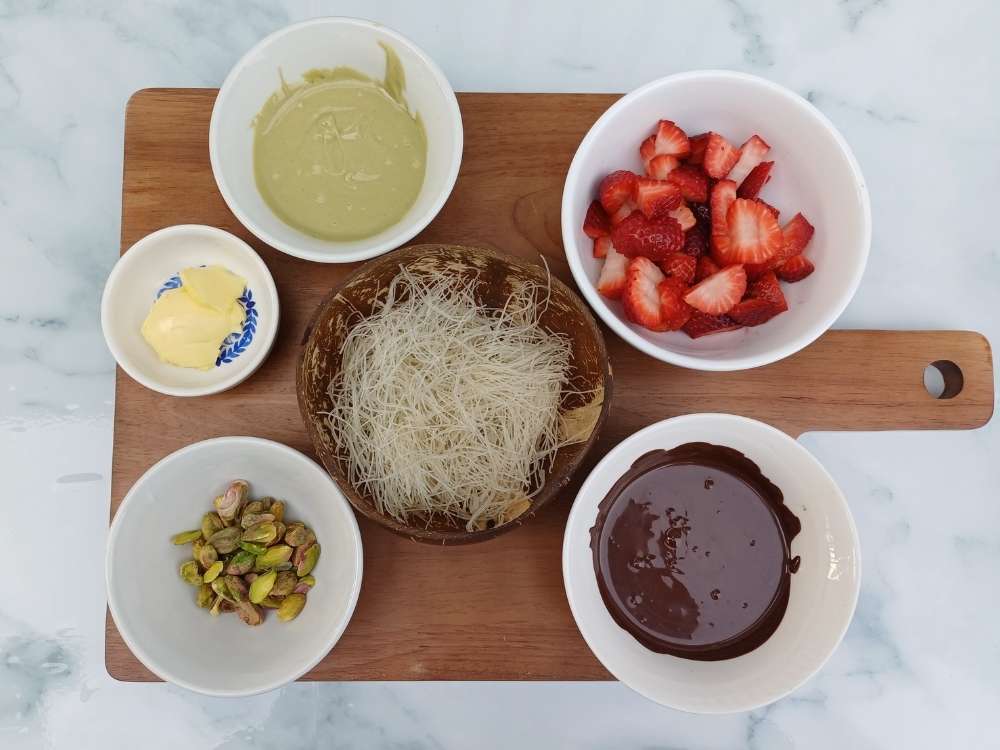 Ingredients for the Dubai chocolate strawberry cup recipe on a board in white bowls including strawberries, pistachio butter, kataifi, pistachios and butter