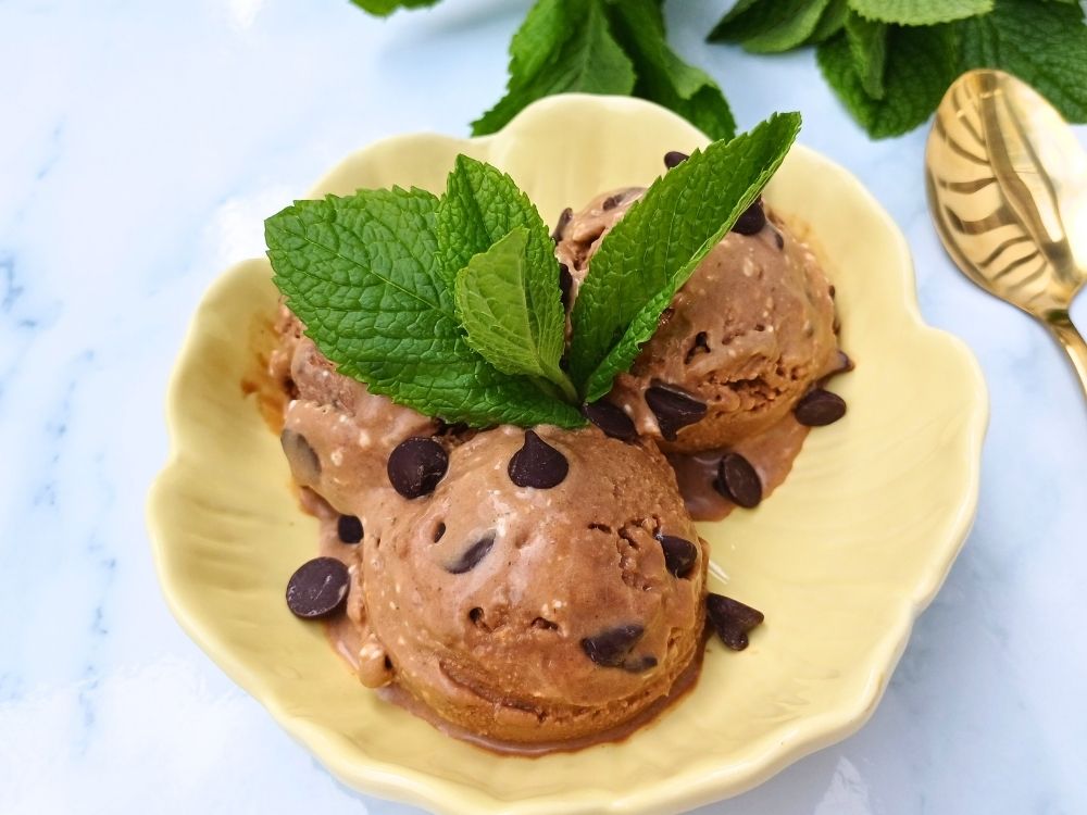 Three scoops on Ninja creami protein ice cream in chocolate flavor with chocolate chips and fresh mint leaves on top in a yellow flower shaped bowl.
