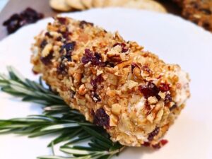 Cranberry pecan goat cheese appetizer log on a white plate with rosemary strings as a decoration under the goat cheese log