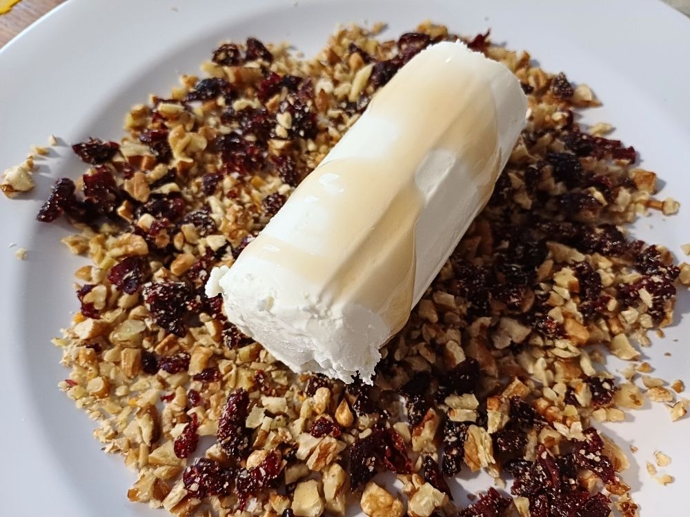 A goat cheese log is on top of chopped pecan nuts and cranberries on a plate.