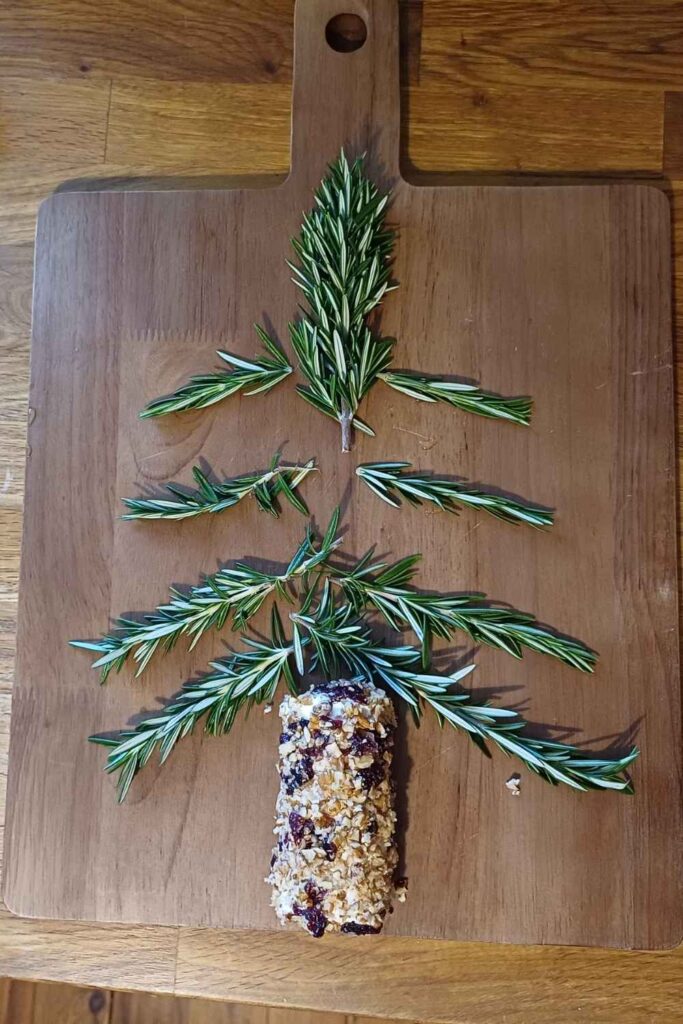 Rosemary springs laid out on a wooden board in a shape of a christmas tree with a goat cheese log at the bottom rolled in pecan nuts for a tree trunk