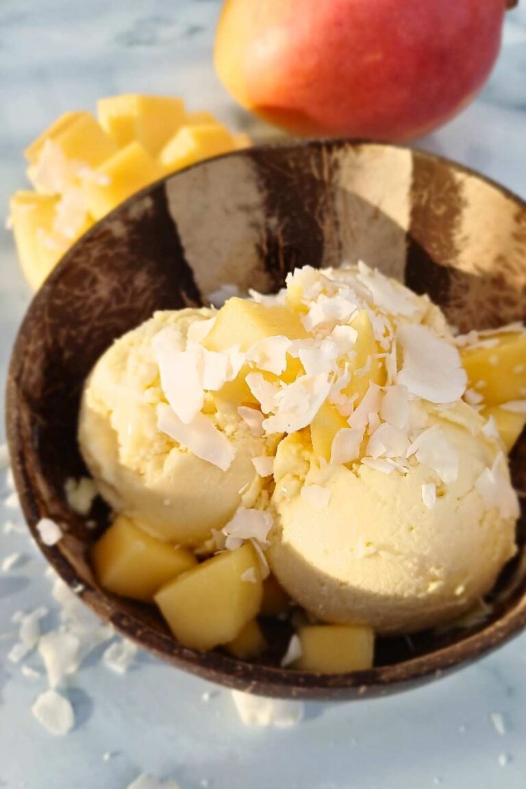 scoops of mango ice cream in a coconut shell bowl with fresh mangos on top and in the background