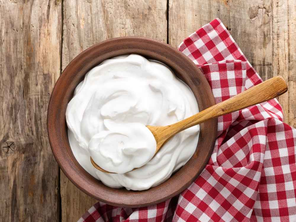 A wooden bowl with fresh Greek Yogurt with a wooden spoon and red and white checkered napkin on a wooden table