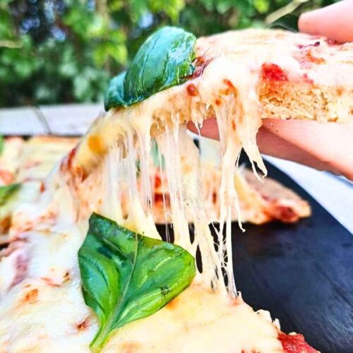 A slice of lmond flour pizza crust with melted cheese and fresh basil being lifted off the board